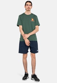 Dark green cotton t-shirt with orange graphic print and crew neckline, paired with dark navy shorts. Black athletic shoes complete the look.