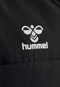 Sort stof har et hvidt Hummel-logo med et bistrefe design over mærkenavnet. Materialet har en glat tekstur, der er velegnet til sportstøj.
