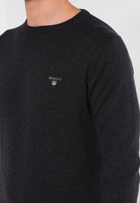 Maglione grigio scuro a girocollo realizzato in morbido tessuto lavorato a maglia. Presenta un piccolo logo GANT ricamato sul petto. Design di taglio classico.