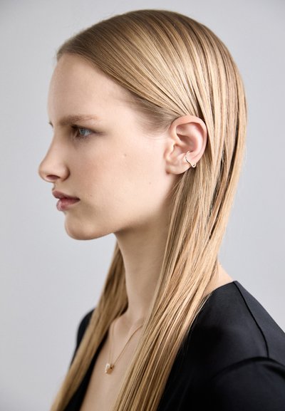Boucle d'oreille en argent présentée sur l'oreille, avec un design ondulé. Le mannequin a de longs cheveux blonds raides et porte un haut noir.