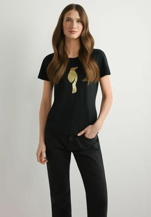 Kobieta z długimi brązowymi włosami, ubrana w czarny t-shirt z złotym motywem ptaka i czarne spodnie, stojąca na tle jasnoszarego tła.