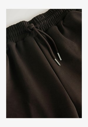 Brune joggers med elastisk talje og snor. Glat tekstur med sølvfarvede spidser på snoren. Ingen synlige lommer.