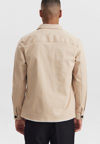 Beige puuvillatakki napillisen kauluksen, pitkien hihojen ja rentona istuvuutena. Ominaisuuksiin kuuluu sileä tekstuuri ja yksi nappiyksityiskohta jokaisessa hihassa.