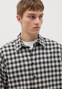 Chemise à carreaux noir et blanc avec un col, design à boutons et texture de tissu douce. Positionnée partiellement sur un mannequin contre un fond uni.