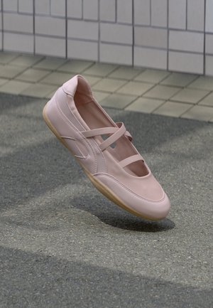 Lichtroze balletflat met elastische kruissriempjes en een beige zool, zwevend boven grijze betontegels met structuur.