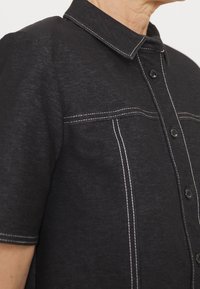 Camicia nera a maniche corte con cuciture a contrasto bianche, bottone frontale e colletto a punta, indossata sulla parte superiore del torso e del collo di una persona.