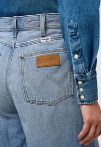 Ljusblå denimjeans med ett läderpatch från Wrangler, med sydda fickor och en knapp-up denimskjorta med pärlknappar.