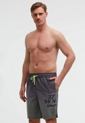 Hemdloser Mann trägt graue Badeshorts mit neongrünem Kordelzug und schwarzem Schriftzug, steht mit einer Hand in der Tasche vor weißem Hintergrund.