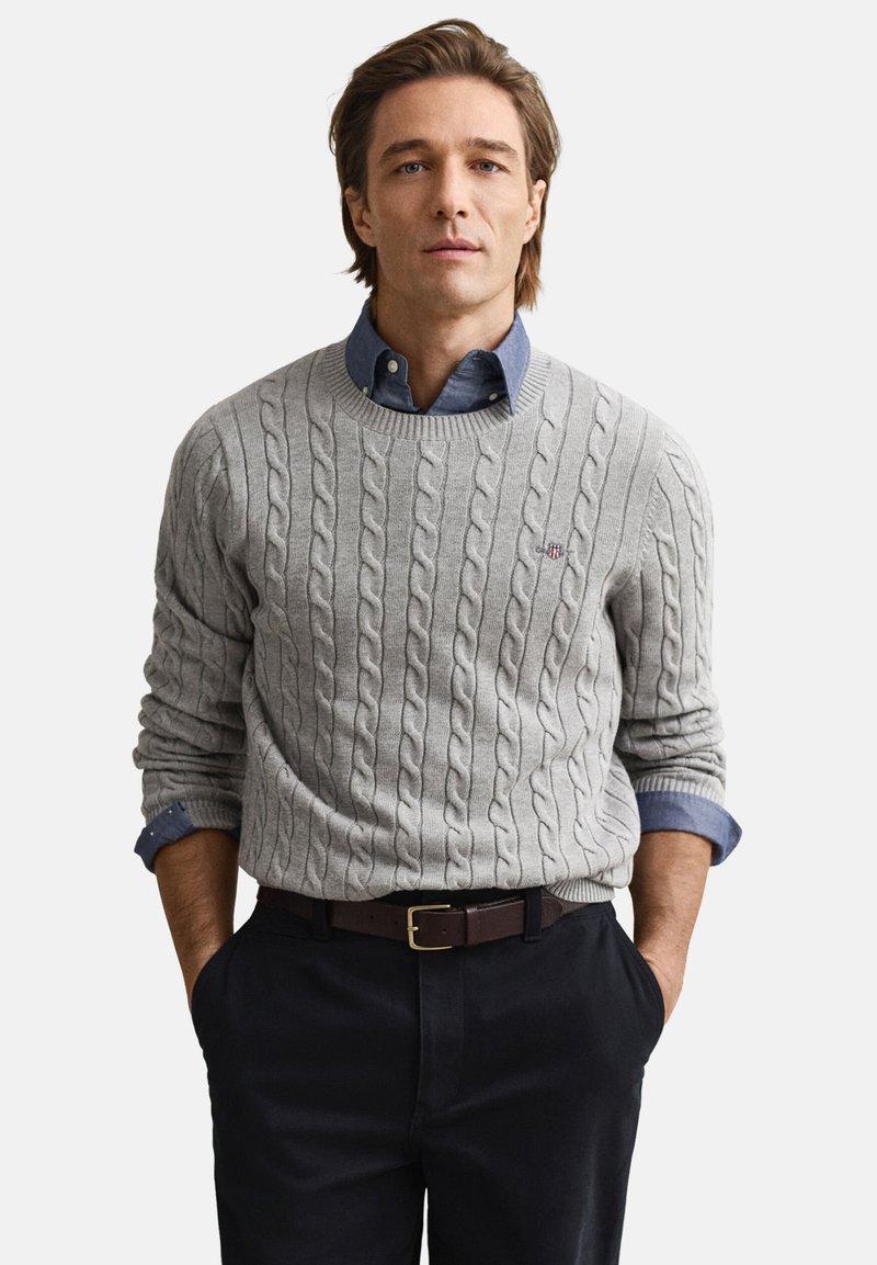 Sivi pulover z vzorcem kabelskega pletenja z okroglim izrezom, oblečen preko modre srajce z ovratnikom. Temne hlače in rjavi pas dopolnjujejo obleko.