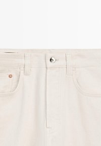 Bund einer cremefarbenen Denim-Jeans mit Gürtelschlaufen, vorderem Knopfverschluss und sichtbaren Vordertaschen.