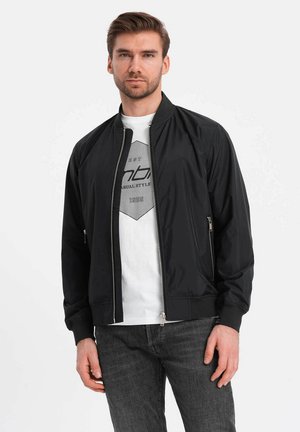 Veste bomber noire avec fermeture éclair sur le devant, poignets côtelés et texture lisse. Portée sur un t-shirt graphique blanc et un jean gris.