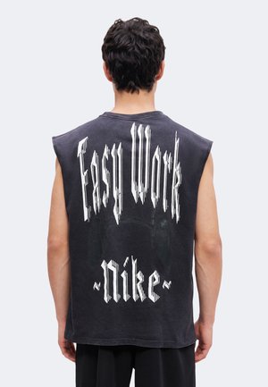 Mężczyzna z ciemnymi kręconymi włosami, ubrany w czarny bezrękawnik z napisem „Easy Work Nike” na plecach, widoczny od tyłu.