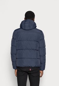 Veste matelassée bleu marine avec une capuche, présentant un design matelassé, une texture lisse et des poignets élastiques, vue de dos.