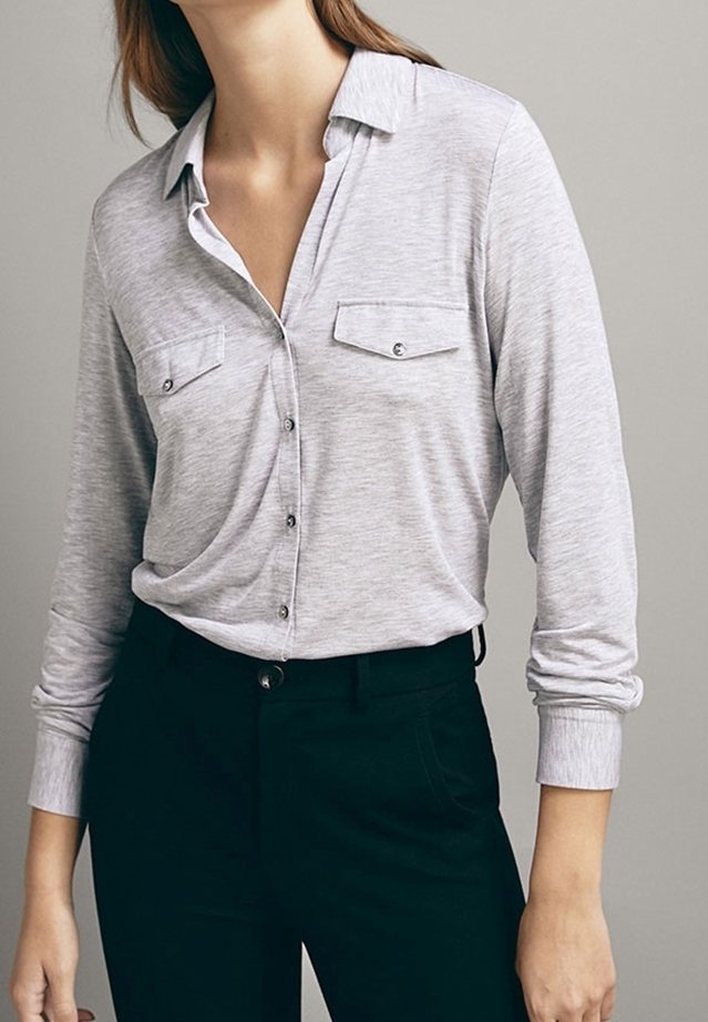 Helle graue Langarmshirt mit einem Button-Down-Kragen, zwei Brusttaschen und einer weichen Textur, kombiniert mit schwarzen, enganliegenden Hosen.