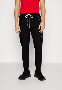 Svarta cargo-byxor med dragkedjefickor, snörning i midjan i vitt, tapered passform, ihop med en röd t-shirt och svarta sneakers.