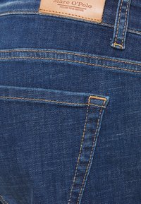 Tejanos de mezclilla azul con una textura suave, que presentan costuras naranjas en contraste y una etiqueta de cuero en la pretina. Bolsillos traseros visibles.