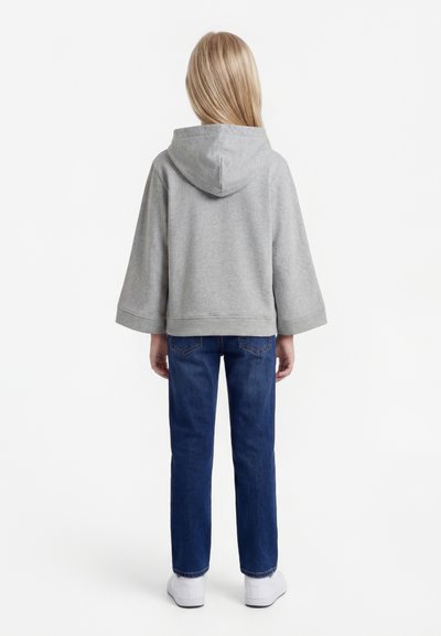 Lapsel, kellel on pikad blondid juuksed, seljas lahtine hall kapuutsiga dressipluus, sinised teksad ja valged tossud, seisab valgel taustal seljaga vaataja poole.