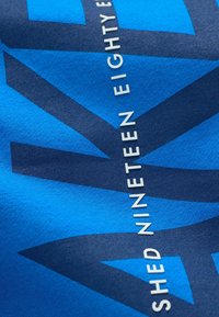 Blauwe textiel met een groot donkerblauw grafisch ontwerp en de tekst "SHEEN NINETEEN EIGHTY" in witte verticale letters.