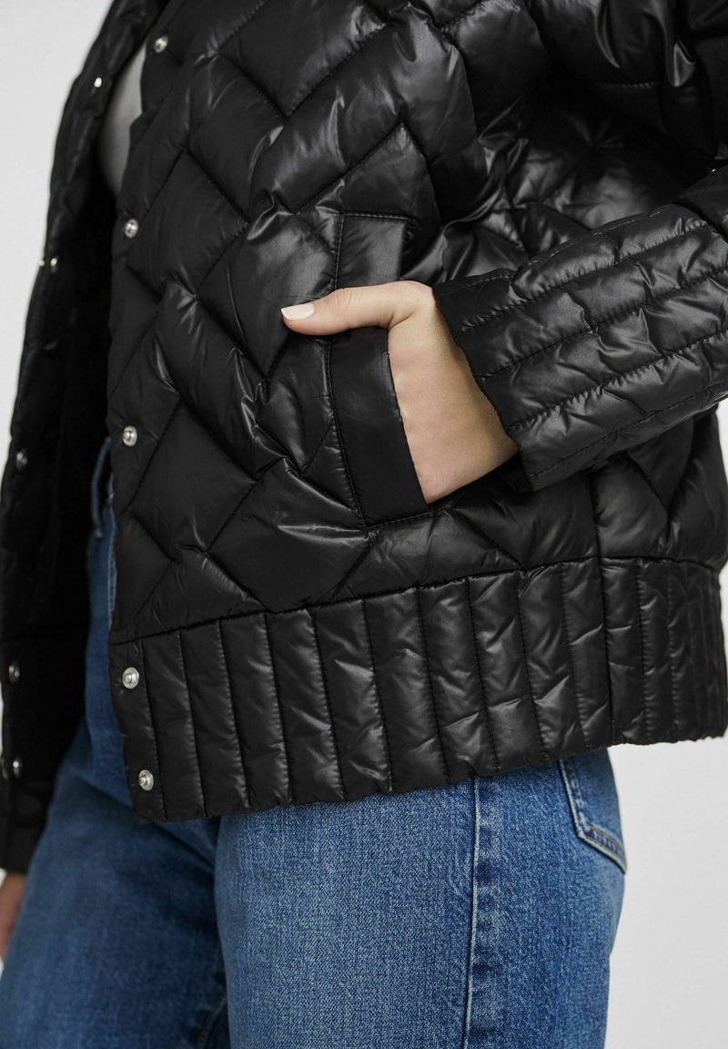 Giubbotto nero imbottito con un design trapuntato, dotato di bottoni a pressione e una tasca laterale. La texture è liscia e lucida, con orlo a coste.