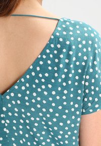 Prenda color teal con un profundo escote en V y mangas cortas, decorada con lunares blancos. Textura suave con un diseño ligero y fluido.