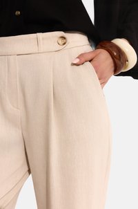 Beżowe spodnie plisowane o gładkiej fakturze, z kieszenią po bokach i złotym zapięciem na guziki w talii.