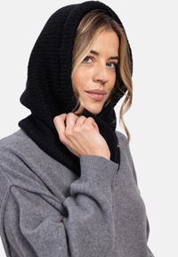 Czarny dzianinowy kaptur zarzucony na szyję, w połączeniu z szarym swetrem o szerokich rękawach i strukturalnej tkaninie. Luźny i warstwowy fason.