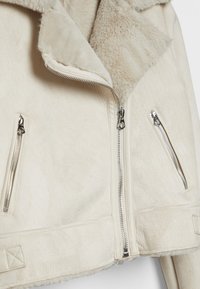 Krémová bunda z umělé kožešiny s širokým límcem, stříbrnými zipy a dvěma zipovými bočními kapsami s texturovaným povrchem.