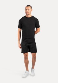Camiseta de deporte negra de manga corta con un diseño ajustado, combinada con pantalones cortos negros y zapatillas blancas, de pie contra un fondo neutro.