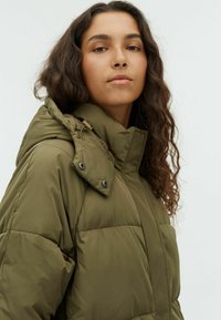 Woman with long curly dark hair wearing an olive green padded jacket with a high collar against a plain white background.