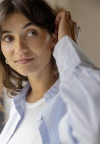 Femme aux yeux marron et aux cheveux courts et bouclés ajuste sa boucle d'oreille, portant une chemise blanche sous une chemise bleu clair à boutons.
