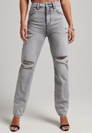 Jean taille haute gris clair avec de grands trous déchirés aux deux genoux, porté avec des talons noirs ouverts et des accessoires argentés.