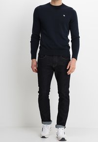 Granatowy sweter dzianinowy z okrągłym dekoltem i prążkowanymi mankietami, zestawiony z ciemnymi dżinsami i białymi trampkami. Na piersi znajduje się małe logo.