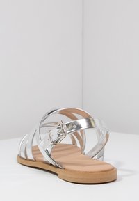 Sandal in metallo argentato con design a strap incrociati, dettaglio fibbia e soletta color tan. Suola piatta con texture liscia e forma arrotondata.