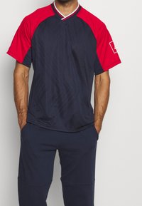 Maillot de sport bleu marine à manches courtes rouges, col en V, et motif texturé. Présente une bordure blanche sur l'encolure et un logo sur la manche.