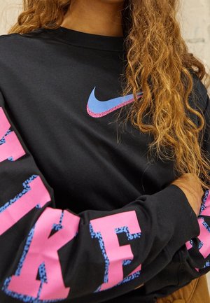 Person with long curly hair wearing black Nike sweatshirt with blue and pink logo on chest and pink text on sleeve.