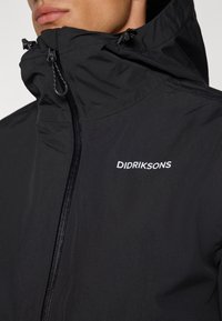 Veste noire imperméable avec un col haut, fermeture éclair et tissu texturé. Présente un logo blanc "DIDRIKSONS" sur la poitrine.