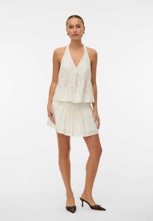 Femme debout portant un haut dos nu blanc à œillets et une jupe assortie, associée à des escarpins marron à bouts pointus.