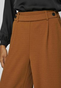 Brown tailored trousers with a smooth texture, featuring a high waist, two front pleats, and black button accents on the waistband.