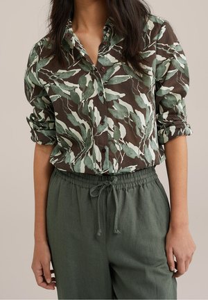 Vrouw draagt een groen-bruine blouse met bladmotief, met opgerolde mouwen en donkergroene broek met trekkoord.
