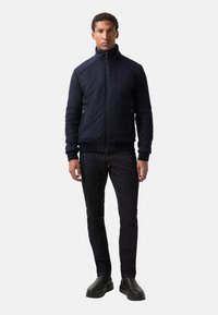 Mann in einer marineblauen Zip-Jacke, dunklen Jeans und schwarzen Stiefeln, der mit entspannten Armen vor einem schlichten weißen Hintergrund steht.