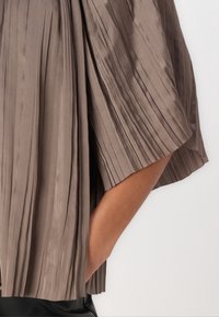 Jupe plissée taupe avec une fente mi-longue révélant une jambe bronzée et une botte en cuir noir.