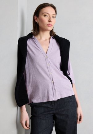 Γυναίκα που στηρίζεται σε τοίχο, φορώντας μωβ πουκάμισο με κουμπιά, με μαύρο πουλόβερ περασμένο στους ώμους και σκούρο παντελόνι.