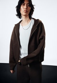 Hoodie zippé marron en tissu doux avec poches kangourou, associé à une chemise bleu clair. Texture lisse, coupe décontractée et design superposé.