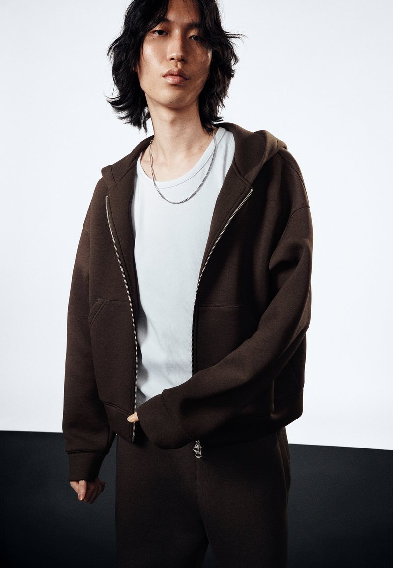 Hoodie zippé marron en tissu doux avec poches kangourou, associé à une chemise bleu clair. Texture lisse, coupe décontractée et design superposé.