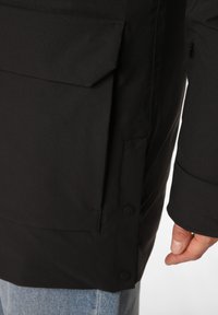 Black waterproof jacket with a side pocket and snap-button closure. Features a smooth texture and a slightly loose fit, visible on the arm.