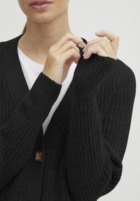 Čierny rebrovaný cardigan s detailom gombíka vpredu. Dlhé rukávy majú štruktúrovanú látku. Nosený cez bielu košeľu.