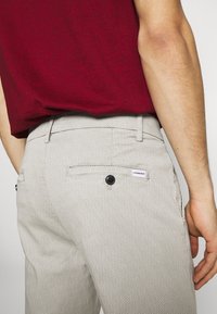 Homme portant un pantalon léger gris à motifs avec une poche arrière à boutons et une chemise rouge, la main reposant sur la hanche.
