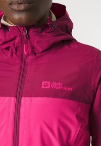 Roze hooded jack met een contrasterend donkerroze paneel. Voorzien van een ritssluiting en een logo op de borst. Gladde afwerking, lichtgewicht materiaal.