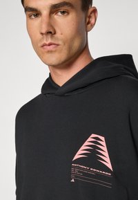 Sudadera negra con un gráfico frontal que presenta formas triangulares rosas y texto. Textura suave con capucha y puños acanalados.