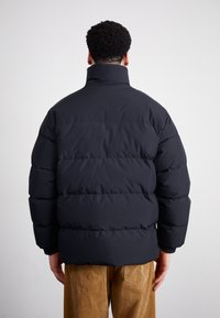 Schwarze Steppjacke mit hohen Kragen, mit einem gesteppten Design und Rippbündchen. Über Cordhosen getragen, Rückansicht gezeigt.